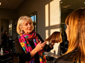 Femme de 58 ans essayant différentes longueurs de cheveux devant un miroir de salon avec une amie, écharpe florale colorée